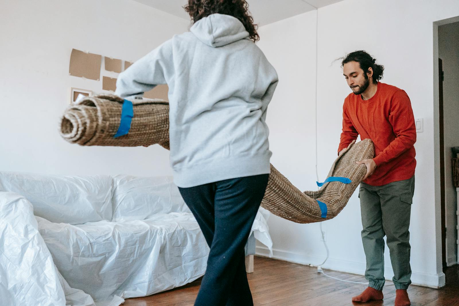 A couple unrolls a carpet in their new apartment, amidst moving and unpacking.